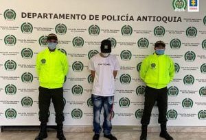 En la fotografía está el procesado y dos policías a los lados usando tapabocas y con un pendón con el escudo de la Policía en mosaico