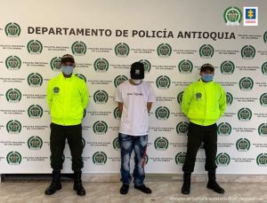 En la fotografía está el procesado y dos policías a los lados usando tapabocas y con un pendón con el escudo de la Policía en mosaico