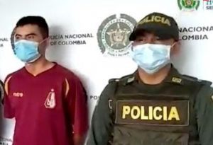 en la imagen se ve una persona detenida bajo custodia de dosuniformados de la Policía Nacional. Detrás suyo un backing institucional.