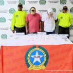 En la fotografía aparecen dos hombres de pie, con las cabezas agachadas, con gorras, se encuentran esposados y custodiados por dos uniformados de la Policía Nacional. Delante de ellos hay una mesa con municiones y estupefacientes.