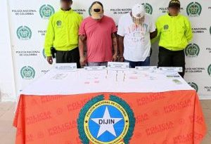 En la fotografía aparecen dos hombres de pie, con las cabezas agachadas, con gorras, se encuentran esposados y custodiados por dos uniformados de la Policía Nacional. Delante de ellos hay una mesa con municiones y estupefacientes.