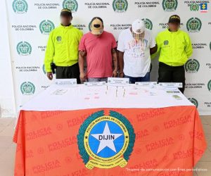 En la fotografía aparecen dos hombres de pie, con las cabezas agachadas, con gorras, se encuentran esposados y custodiados por dos uniformados de la Policía Nacional. Delante de ellos hay una mesa con municiones y estupefacientes.