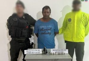 En la fotografía aparece un hombre de mediana edad y estatura, moreno, de pie, con los brazos atrás, vestido con una camiseta azul, custodiado por dos uniformados de la Policía Nacional. Delante de ellos hay una mesa con el arma y las municiones incautadas.