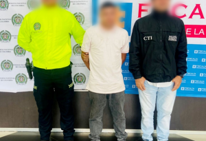 Se visualiza al capturado junto a un uniformado de la Policía Nacional y un investigador del CTI. Detrás de ellos se ubica los banners que identifican a la Policía Nacional y a la Fiscalía