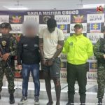 El capturado viste camiseta blanca y pantalón corto negro, está esposado y de frente.  A un costado lo custodia un funcionario del Cuerpo Técnico de Investigación (CTI) y otro del Ejército Nacional.  Al otro costado uno de la Policía Nacional y otro del Ejército.  Atrás, un pendón de la Fiscalía General de la Nación y el Ejército.