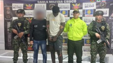 El capturado viste camiseta blanca y pantalón corto negro, está esposado y de frente.  A un costado lo custodia un funcionario del Cuerpo Técnico de Investigación (CTI) y otro del Ejército Nacional.  Al otro costado uno de la Policía Nacional y otro del Ejército.  Atrás, un pendón de la Fiscalía General de la Nación y el Ejército.