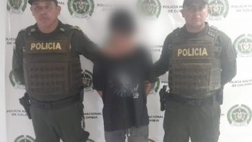 capturado de frente custodiado por dos policías, detrás bannerde la Policía Nacional.