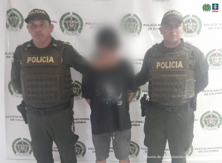 capturado de frente custodiado por dos policías, detrás bannerde la Policía Nacional.