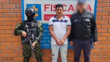 El capturado se encuentra de frente  y custodiado por un servidor del Cuerpo Técnico de Investigación CTI y un uniformado del Ejército Nacional. Detrás de la foto hay un backing de la Fiscalía General de La Nación.