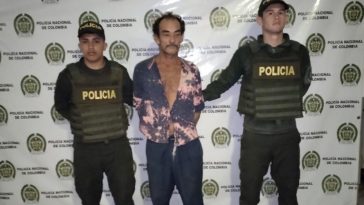 capturado de frente custodiado por dos policías. Detrás banner de la Policía Nacional.