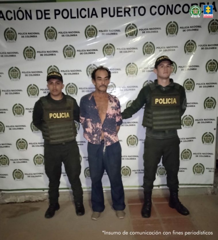 capturado de frente custodiado por dos policías. Detrás banner de la Policía Nacional.