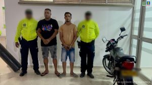 En la fotografía aparecen dos hombres de pie, con los brazos adelante, esposados, ambos vestidos con bermudas de jean y camisetas. Se encuentran custodiado por dos uniformados de la Policía Nacional.