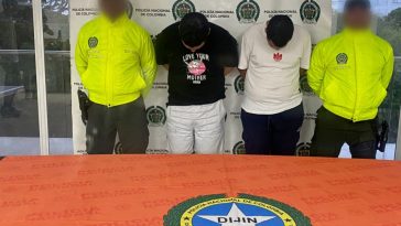 En la fotografía están dos hombres de pie, con las cabezas agachadas, los brazos atrás, custodiado por dos uniformados de la Policía Nacional. Detrás de ellos hay un pendón de esa institución.