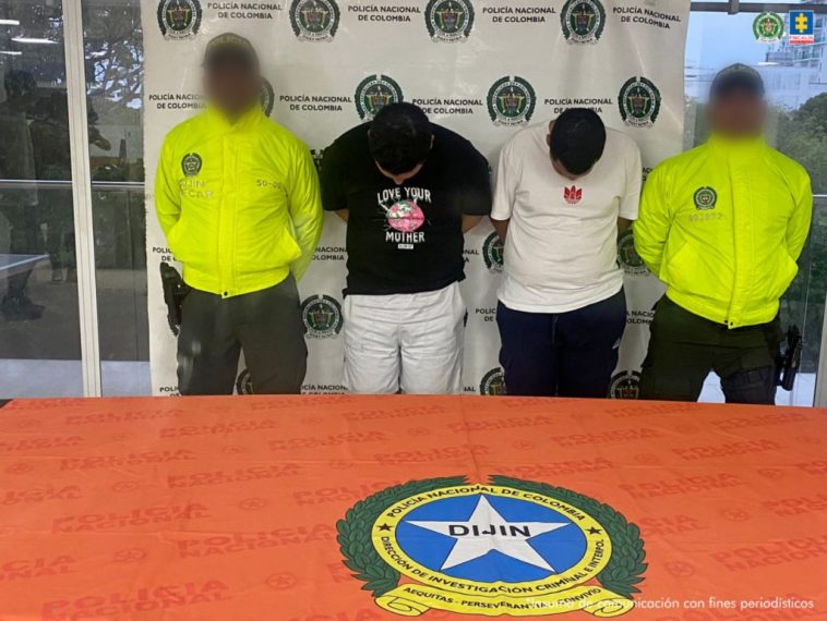 En la fotografía están dos hombres de pie, con las cabezas agachadas, los brazos atrás, custodiado por dos uniformados de la Policía Nacional. Detrás de ellos hay un pendón de esa institución.