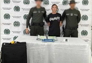 En la imagen está un hombre de tez blanca, de pie, con los brazos atrás, de mediana estatura, vestido con camiseta negra, custodiado por dos agentes de la Policía Nacional. Delante de ellos hay una mesa donde se encuentra las dos bolsas de cocaína.