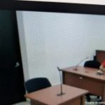 En la imagen está un hombre moreno, de cabellos negros, sentado frente a un escritor de madera con micrófono, al interior de la sala de audiencia se le procesó.  Se encuentra vestido con una camiseta roja y un pantalón azul.