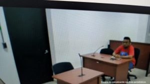 En la imagen está un hombre moreno, de cabellos negros, sentado frente a un escritor de madera con micrófono, al interior de la sala de audiencia se le procesó.  Se encuentra vestido con una camiseta roja y un pantalón azul.