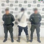 El capturado se encuentra de espaldas y custodiado por dos uniformados de la Policía Nacional. Detrás de la foto hay un backing de la Policía Nacional y el capturado tiene un buzo blanco, jeans y botas.