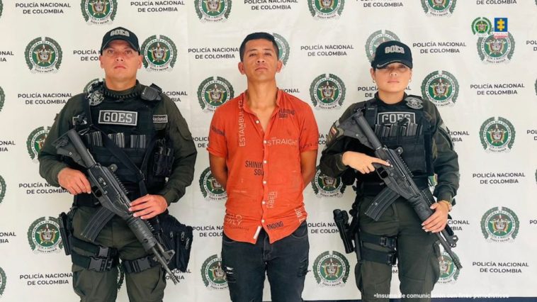 El capturado se encuentra en custodia por dos uniformados del grupo GOES de la Policía Nacional. Detrás se observa un backing de la Policía.