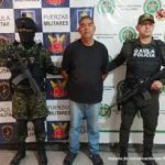 El capturado se encuentra de frente y custodiado por  un uniformado de la Policía y Ejército Nacional. Detrás hay un backing de la Policía y Ejército. El capturado tiene una camiseta de color negro  jeans y tenis.