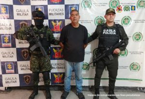 El capturado se encuentra de frente y custodiado por  un uniformado de la Policía y Ejército Nacional. Detrás hay un backing de la Policía y Ejército. El capturado tiene una camiseta de color negro  jeans y tenis.