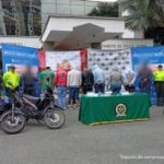 Se observan en fila siete personas capturadas, todos de espaldas y en fila. A cada lado los custodian funcionarios del CTI, Policía Nacional y Ejército Nacional. Al frente de la fila se observan 4 motocicletas incautadas y en una mesa el material incautado