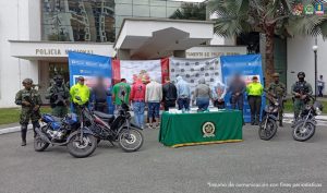 Se observan en fila siete personas capturadas, todos de espaldas y en fila. A cada lado los custodian funcionarios del CTI, Policía Nacional y Ejército Nacional. Al frente de la fila se observan 4 motocicletas incautadas y en una mesa el material incautado