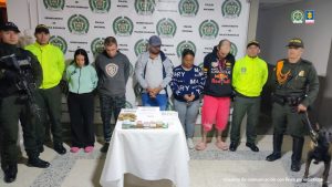 Se observan CINCO personas en fila y de frente, A cada costado funcionarios de Policía Nacional, en uno de los costados un canino de la Policía Nacional.  Atrás un pendón de Policía NAcional, al frente una mesa con elementos incautados