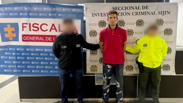 Se visualiza al capturado junto a un investigador del CTI y un uniformado de la Policía Nacional. En la parte posterior los banners institucionales de la Fiscalía General de la Nación y de la Policía Nacional