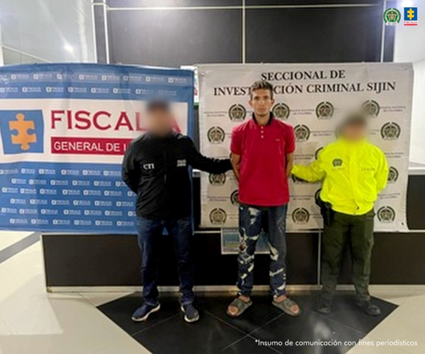 Se visualiza al capturado junto a un investigador del CTI y un uniformado de la Policía Nacional. En la parte posterior los banners institucionales de la Fiscalía General de la Nación y de la Policía Nacional