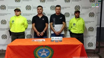 en la imagen se ven dos personas detenidas bajo custodia de integrantes de la Policía Nacional. Detrás suyo un backing institucional. Frente a ellos una mesa con elementos incautados.