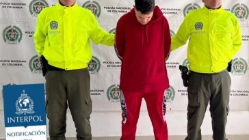 Se observa al capturado de frente con la cabeza abajo, viste chaqueta y pantalón rojo, zapatos tenis blancos.  A cada lado lo custodia un uniformado de la Policía Nacional.  Atrás, un pendón de Policía Nacional y sobrepuesto un texto que dice INTERPOL NOTIFICACIÓN AZUL.