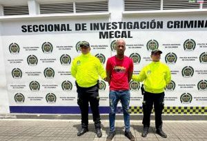 En la fotografía se aprecia de pie y con los brazos atrás a Luis Alfredo Mina Caicedo. Viste camiseta de manga corta de color rojo cn estampado en el pecho negro y amarillo; jean azul desgastado y zapatos negros. A sus extremos hay dos uniformados de la Policía Nacional y detrás de ellos hay un pendón institucional de la Policía.