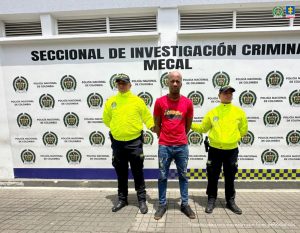 En la fotografía se aprecia de pie y con los brazos atrás a Luis Alfredo Mina Caicedo. Viste camiseta de manga corta de color rojo cn estampado en el pecho negro y amarillo; jean azul desgastado y zapatos negros. A sus extremos hay dos uniformados de la Policía Nacional y detrás de ellos hay un pendón institucional de la Policía.