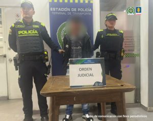 en la imagen se ve una persona detenida bajo custodia de dos integrantes de la Policía Nacional. Detrás suyo un backing institucional.