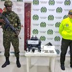 En la imagen se ve una mesa con un dron y otros elementos incautados bajo custodia de un militar y un policial. Detrás suyo backings institucionales.