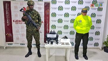 En la imagen se ve una mesa con un dron y otros elementos incautados bajo custodia de un militar y un policial. Detrás suyo backings institucionales.