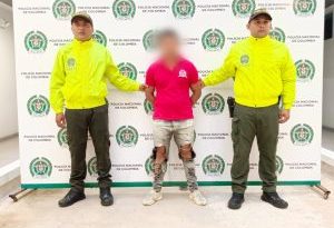 en la imagen se ve una persona detenida bajo custodia de dos uniformados de la Policía Nacional. Detrás suyo un backing institucional.