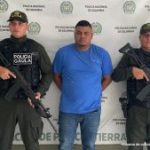 En la fotografía está un hombre de pie, de contextura mediana, moreno, de cabellos cortos crespos, con los brazos atrás esposado. Se encuentra vestido con una camiseta azul un jean azul y custodiado por dos uniformados de la Policía Nacional.