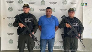 En la fotografía está un hombre de pie, de contextura mediana, moreno, de cabellos cortos crespos, con los brazos atrás esposado. Se encuentra vestido con una camiseta azul un jean azul y custodiado por dos uniformados de la Policía Nacional.