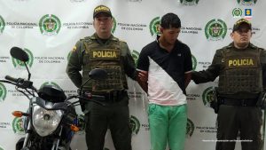 capturado de frente custodiado por dos policías. Al lado, motocicleta incautada. Detrás banner de la Policía Nacional.