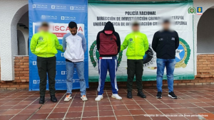 Se visualiza a los capturados junto a uniformados de la Policía Nacional e investigador del CTI. Detrás los banners que identifican a la Fiscalía General de la Nación y a la Policía Nacional. --