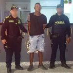 En la imagen se encuentra un hombre de contextura gruesa de pie, con los brazos atrás esposado, vestido con una camiseta negra, bermuda de jean azul y tenis negros. Está custodiado por dos uniformados de la Policía Nacional.