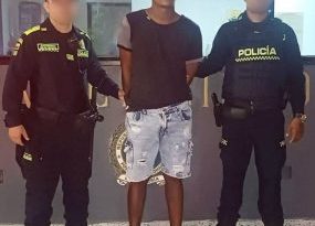 En la imagen se encuentra un hombre de contextura gruesa de pie, con los brazos atrás esposado, vestido con una camiseta negra, bermuda de jean azul y tenis negros. Está custodiado por dos uniformados de la Policía Nacional.