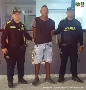 En la imagen se encuentra un hombre de contextura gruesa de pie, con los brazos atrás esposado, vestido con una camiseta negra, bermuda de jean azul y tenis negros. Está custodiado por dos uniformados de la Policía Nacional.