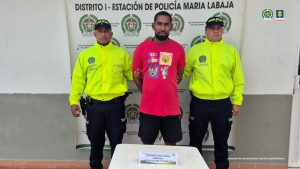 En la fotografía está un hombre de pie, con los brazos atrás esposados, moreno, de cabellos negros, crespos, con barba, vestido con una camiseta roja y una bermuda negra. Se encuentra custodiado por dos uniformados de la Policía Nacional.