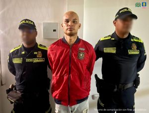 En la foto aparece un hombre trigueño, de pie, con los brazos atrás esposado, cabellos crespos, color amarillo. Se encuentra vestido con una chaqueta roja y jean azul claro. Está custodiado por dos uniformados de la Policía Nacional.