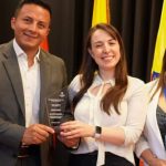 El secretario de gobierno de Sandoná, John Jairo Castillo, y la comisaria de familia, Diana Fernanda Delgado Pabón, reciben un reconocimiento por buenas prácticas de manos de una representante del Ministerio de Justicia y del Derecho, en un evento que destaca el trabajo de varias comisarías de familia en Colombia. Los tres posan sonrientes con el premio, con banderas de Colombia al fondo