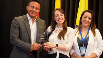 El secretario de gobierno de Sandoná, John Jairo Castillo, y la comisaria de familia, Diana Fernanda Delgado Pabón, reciben un reconocimiento por buenas prácticas de manos de una representante del Ministerio de Justicia y del Derecho, en un evento que destaca el trabajo de varias comisarías de familia en Colombia. Los tres posan sonrientes con el premio, con banderas de Colombia al fondo