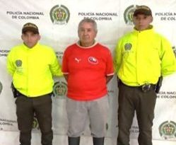 El procesado aparece vestido con una camiseta roja y un pantalón gris. De fondo está el backing de la Policía y dos uniformados.